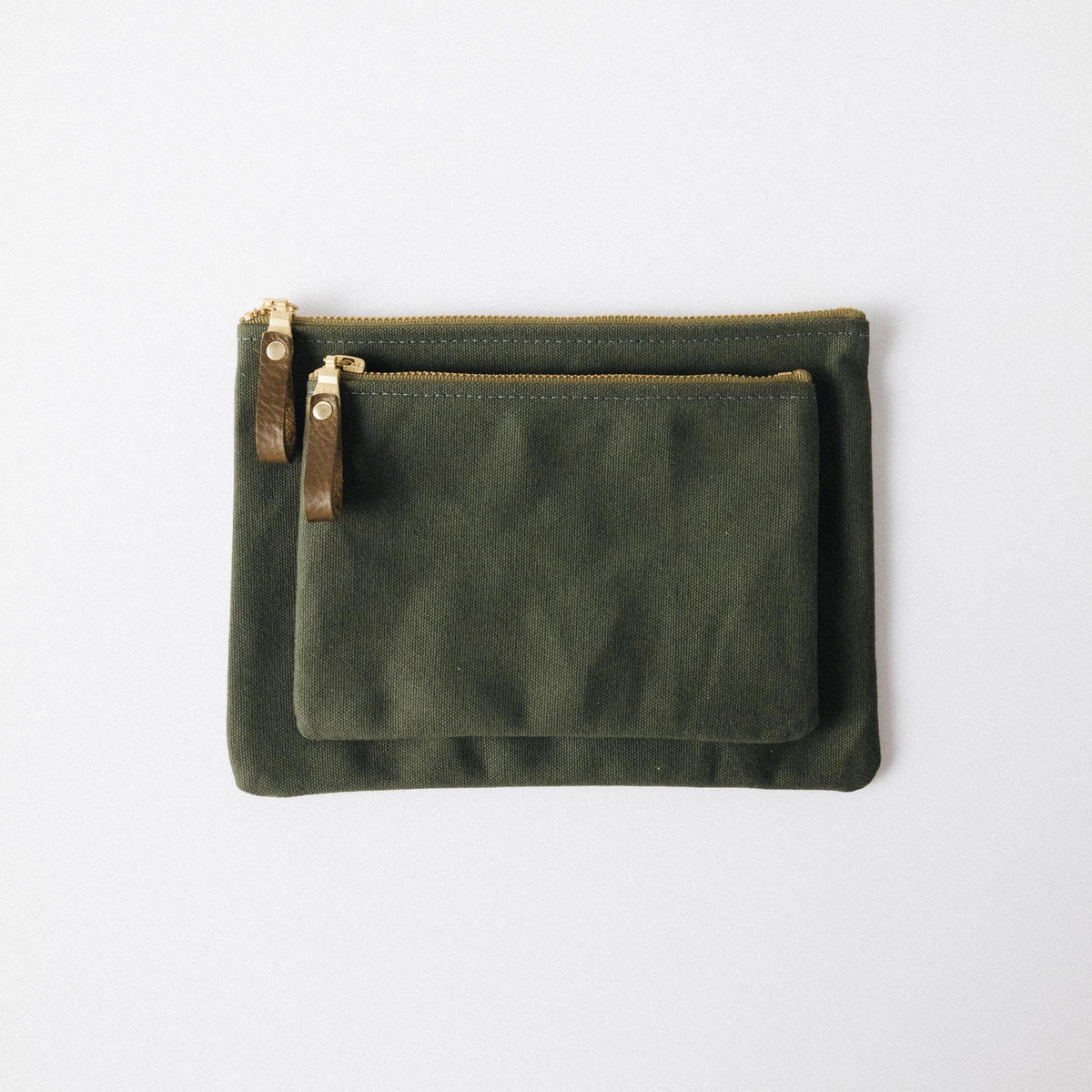 Two olive green cotton canvas zip pouches with solid brass zippers and leather pulls on a white background.