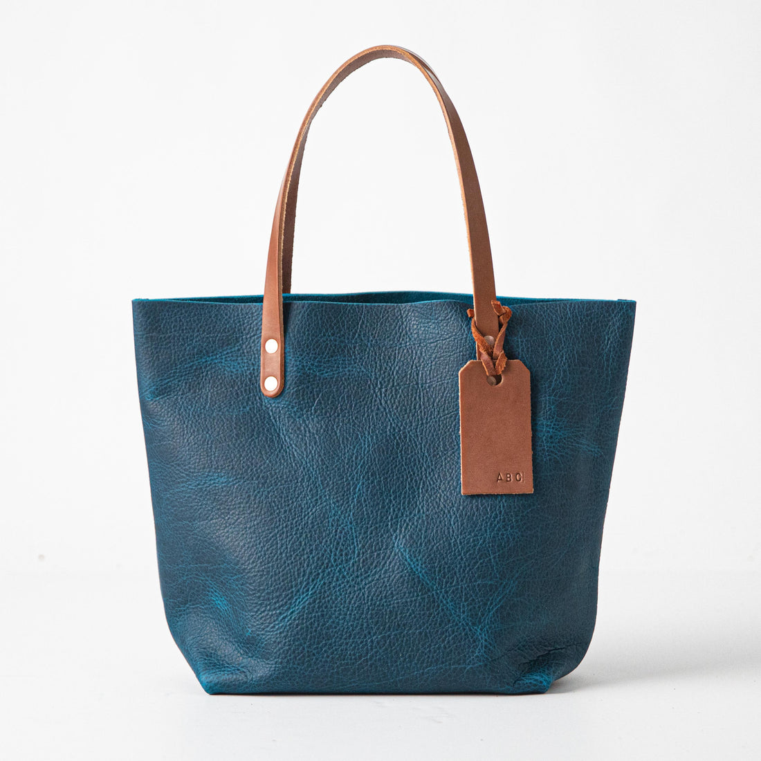 Handmade petrol blue bison leather tote bag with tan handles and a personalized embossed leather luggage tag.
