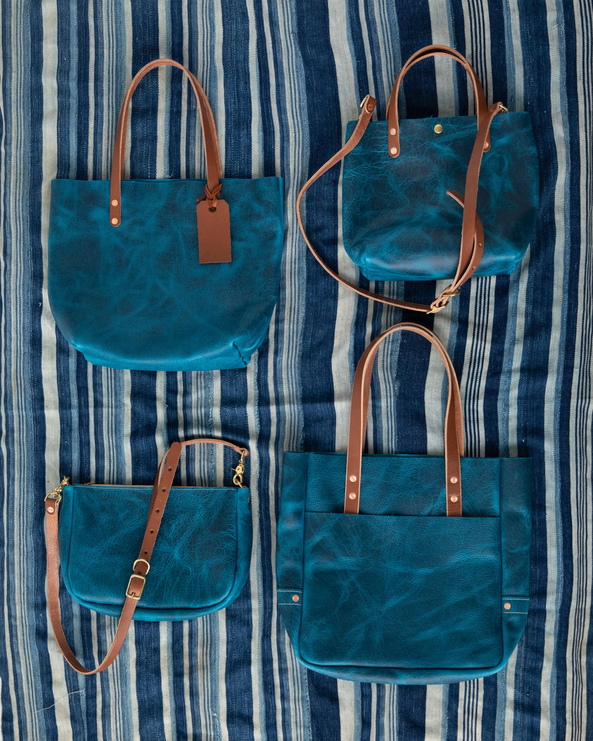 Four Petrol Blue Bison full-grain leather bags with brown straps on a striped blue fabric.