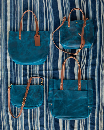 Four Petrol Blue Bison full-grain leather bags with brown straps on a striped blue fabric.