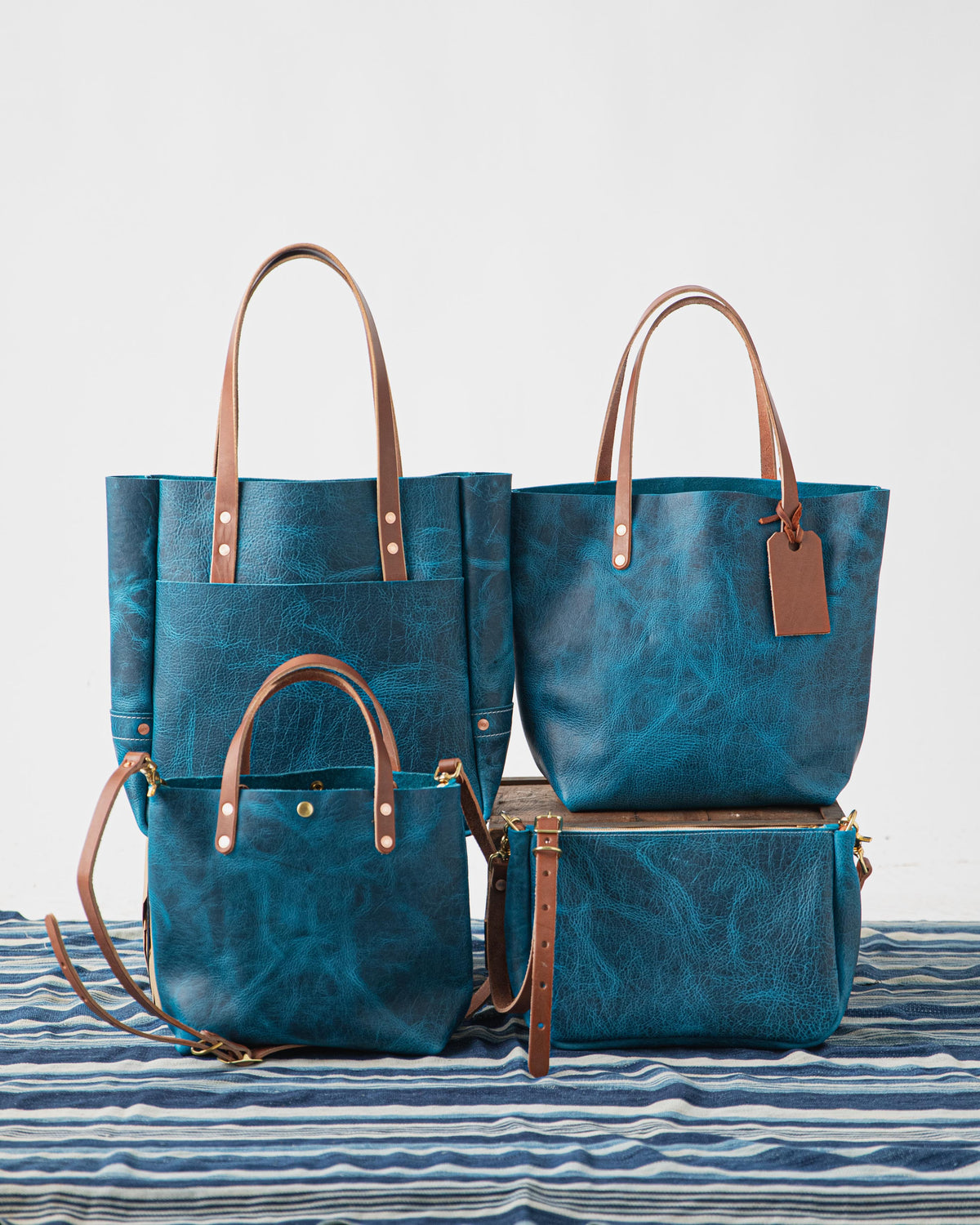 Four Petrol Blue Bison full-grain leather bags: two large totes, a crossbody, and a clutch, with brass hardware and tan straps.