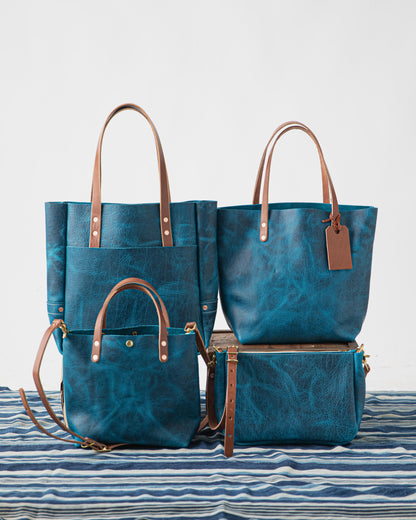 Four Petrol Blue Bison full-grain leather bags: two large totes, a crossbody, and a clutch, with brass hardware and tan straps.