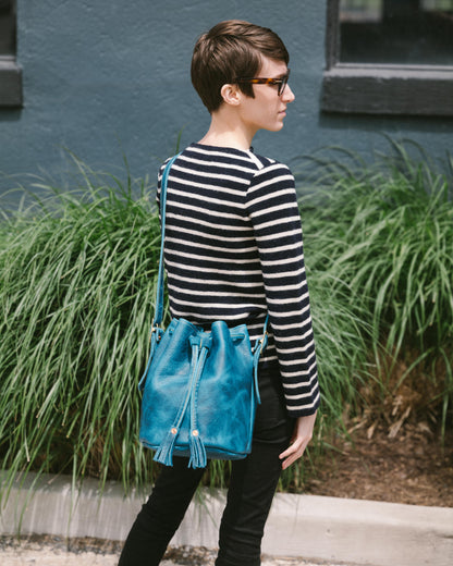 Petrol Blue Bison Bucket Bag with elegant leather tassels, worn crossbody by a person in a striped sweater.